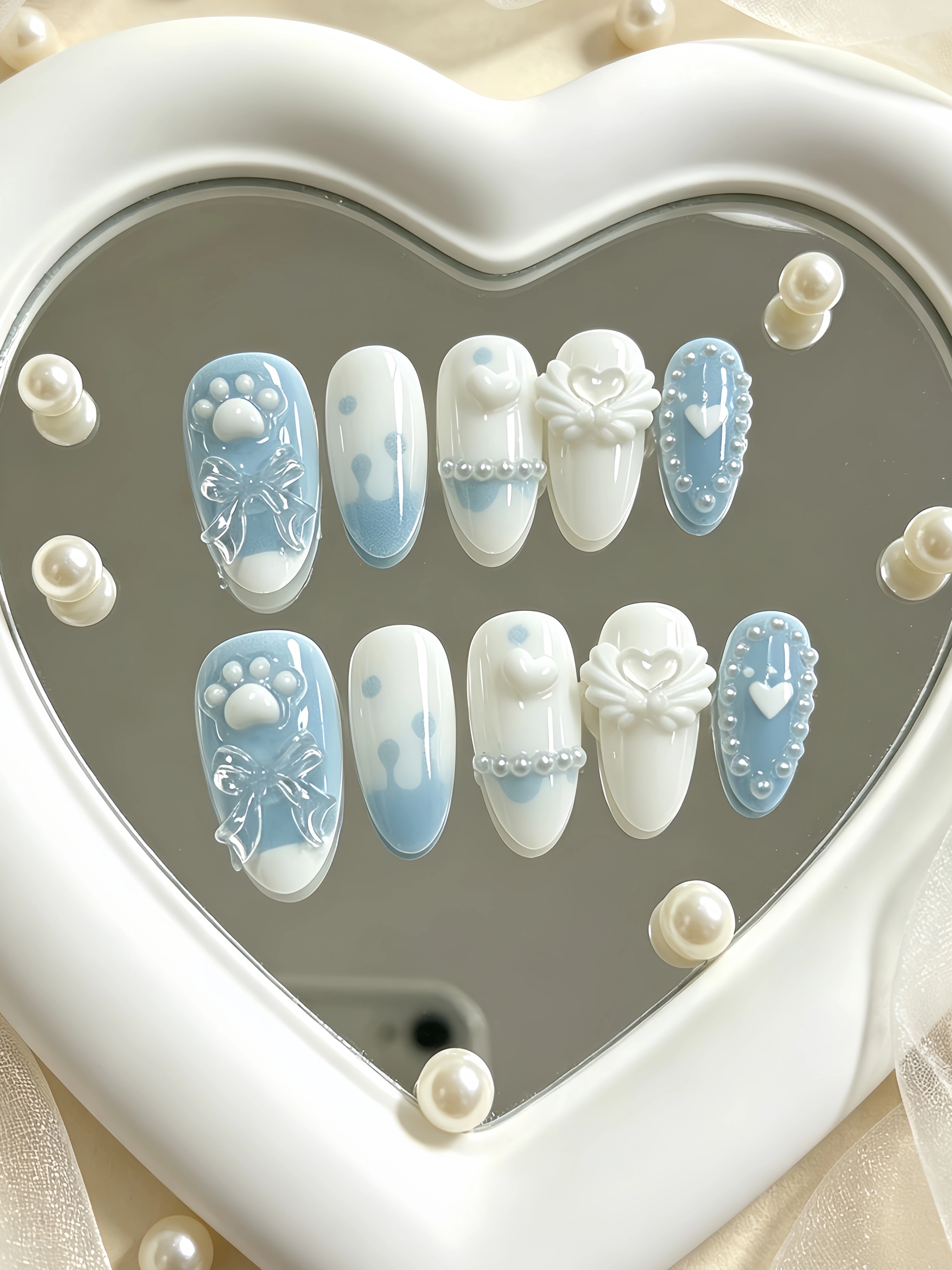 10 ongles de longueur moyenne en forme d'amande, aux couleurs principales blanc laiteux et bleu clair, décorés de nœuds transparents blanc laiteux en forme de patte de chat, d'ailes en forme de cœur,