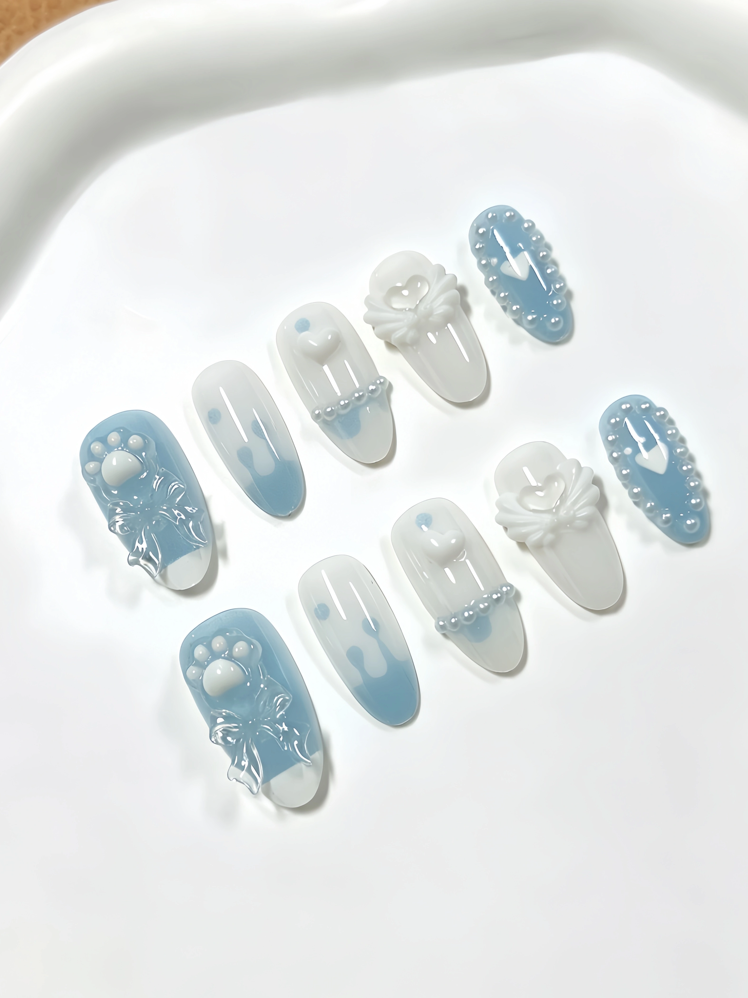 10 ongles de longueur moyenne en forme d'amande, aux couleurs principales blanc laiteux et bleu clair, décorés de nœuds transparents blanc laiteux en forme de patte de chat, d'ailes en forme de cœur,