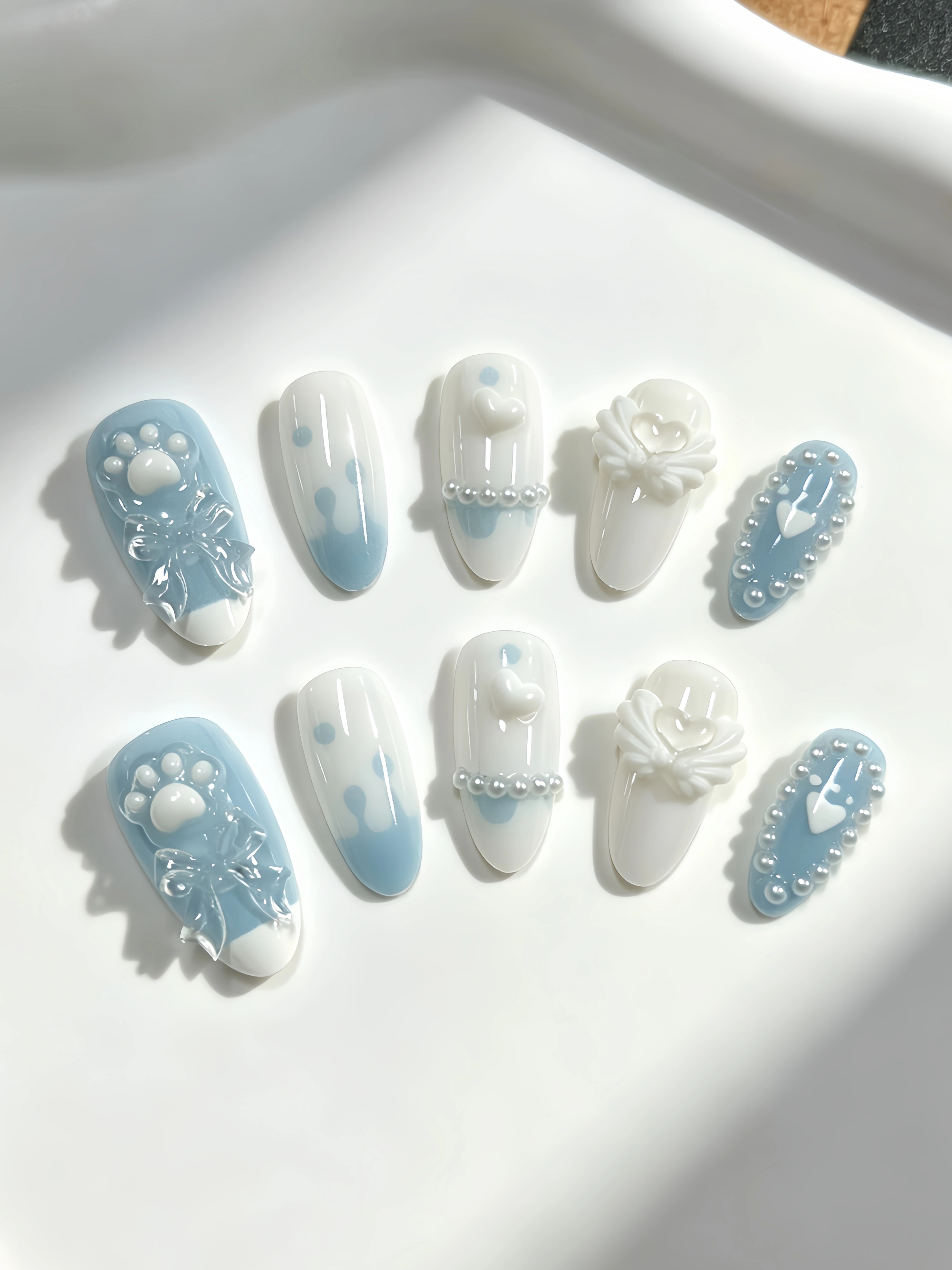 10 ongles de longueur moyenne en forme d'amande, aux couleurs principales blanc laiteux et bleu clair, décorés de nœuds transparents blanc laiteux en forme de patte de chat, d'ailes en forme de cœur,